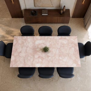 Peut inclure: Une table à manger rectangulaire avec une surface en pierre texturée rose clair, ornée d'une petite plante verte au centre. La table est entourée de huit chaises gris anthracite foncé avec de fins pieds en métal doré, disposées symétriquement.