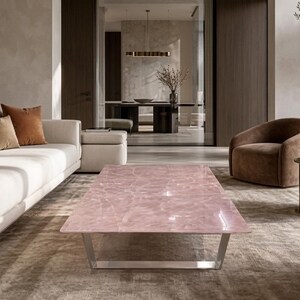 Peut inclure: Une table basse rectangulaire dotée d'un plateau en dalle de quartz rose translucide et poli avec des motifs de veines naturels, reposant sur une base en cadre métallique argenté, placée au centre d'un salon.