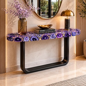 May include: A console table featuring an oval top made of purple agate slices with gold-toned edges, supported by a black U-shaped metal base. The table holds a brown glass vase with purple flowers, a gold bowl, and two books titled Chanel and Vanity Fair.