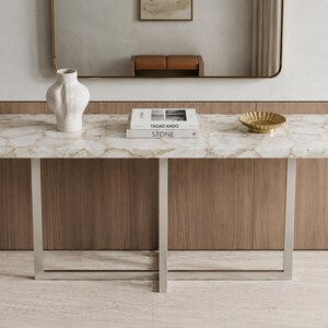May include: A rectangular console table with a white and beige marbled stone top and a silver metal frame. On the table sits a white ceramic vase, two stacked books titled Tadao Ando Stone, and a small gold-toned scalloped bowl.