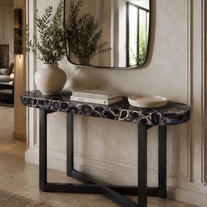 May include: A console table with a black agate stone top featuring white crystalline patterns and a black metal base with an X-shaped support. On the table sits a textured beige vase with green branches, two stacked books, and a shallow beige bowl.