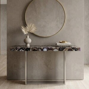 May include: A rectangular console table featuring a polished agate stone top with purple, black, and white banding, supported by a silver-toned metal frame. A beige ceramic vase with dried palm leaves and a stack of books sit on the surface.