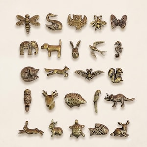 May include: A collection of twenty-five small, antique brass-toned metal animal figurines arranged in five rows. The set includes various creatures such as a dragonfly, swan, owl, bee, butterfly, elephant, dog, rabbit, bird, seahorse, fox, bat, squirrel, deer, hedgehog, parrot, dinosaur, turtle, and fish.