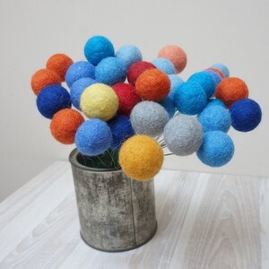 May include: A bouquet of colourful felt balls in shades of blue, orange, yellow, and red arranged in a metal tin.