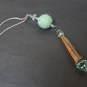 Könnte beinhalten: Eine hellgrüne Filzkugel mit blauen Perlen hängt an einer silbernen Kette und einem langen, klaren Kristallprisma mit braunen Akzenten.