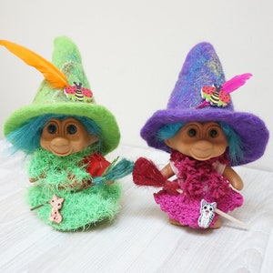 May include: Two troll dolls dressed as witches with green and purple felt hats, blue hair, and colorful dresses. Each doll holds a broom and has a small cat charm on their dress.
