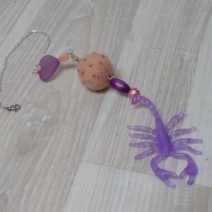 May include: A purple scorpion charm with a pink and white beaded ball and a purple heart charm attached to a silver chain.