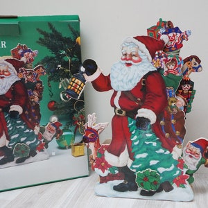 Op de afbeelding: Een rode en witte kerstmanfiguur die een zak met cadeaus en een lantaarn vasthoudt. Hij is omringd door een kerstboom, rendieren en andere feestelijke decoraties. Het figuur is gemaakt van karton en maakt deel uit van een muzikale kerstkaart.