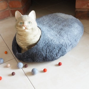 May include: A gray felt cat bed with a ceramic cat figurine inside. The bed is on a white tiled floor with several small, colorful balls scattered around it.
