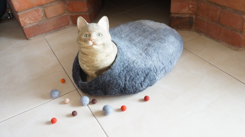 May include: A gray felt cat bed with a ceramic cat figurine inside. The bed is on a white tiled floor with several small, colorful balls scattered around it.