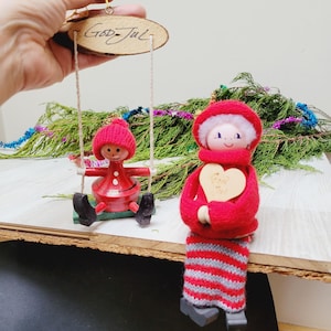Op de afbeelding: Twee kerstversieringen: een rode pop op een schommel en een zittende pop. Het schommelornament heeft een houten basis met de tekst "God Jul". De zittende pop draagt een rode outfit met een houten hartaccent.