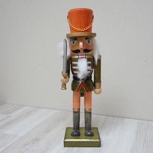 May include: A wooden nutcracker soldier figurine with a green and red uniform, a white beard, and a red and orange hat. The nutcracker is holding a silver sword.