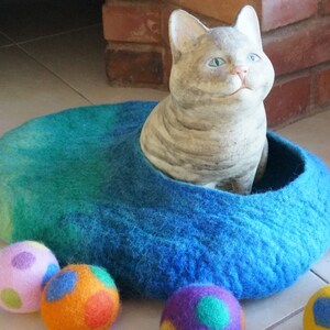 May include: A blue felt cat bed with a gray cat figurine inside. There are five colorful felt balls scattered around the bed.