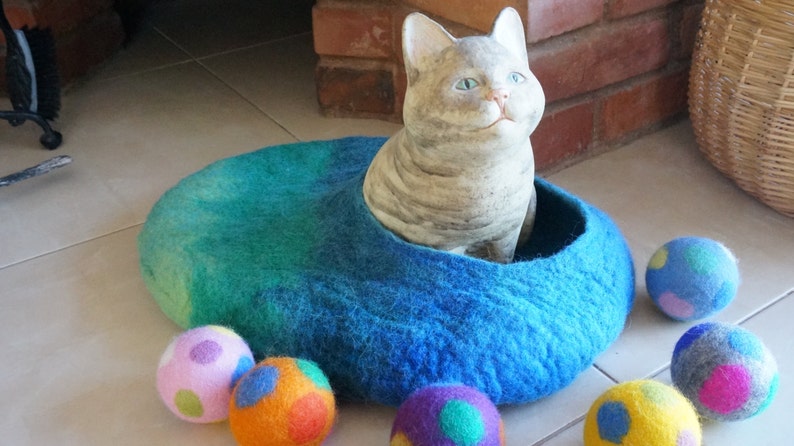 May include: A blue felt cat bed with a gray cat figurine inside. There are five colorful felt balls scattered around the bed.