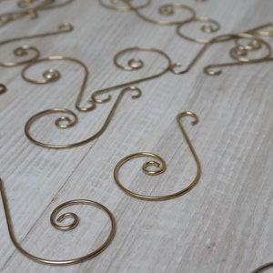May include: A group of gold-colored metal ornament hooks with a decorative swirl design. The hooks are scattered on a light wood surface.