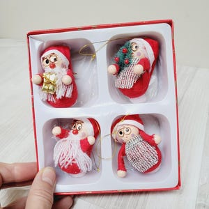 Op de afbeelding: Set van vier houten kerstversieringen in een rode doos. Elke versiering is een klein, rood gekleed figuurtje met een witte baard en een witte hoed. De versieringen zitten in een witte doos met een rode rand.