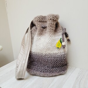 May include: A brown, white, and gray knitted drawstring bag with a long strap. The bag has a small yellow pear-shaped embellishment.