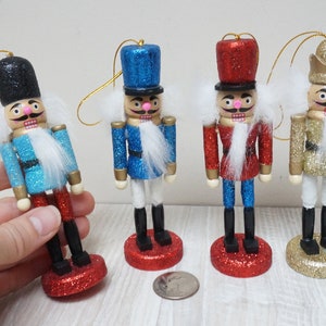 Könnte beinhalten: Vier Nussknacker-Anhänger aus Holz mit Glitzer. Die Nussknacker tragen blaue, rote und goldene Uniformen mit weißem Pelzbesatz. Sie haben schwarze Hüte und rosa Wangen. Die Anhänger hängen an goldenen Schnüren.