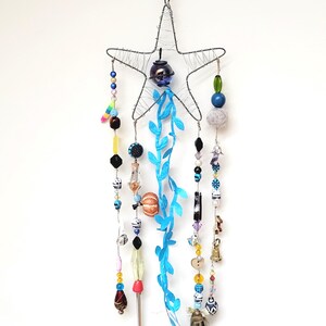 Op de afbeelding: Een handgemaakte stervormige windgong met een draadframe. Het heeft verschillende kleurrijke kralen, een kleine bel en blauwe bladvormige accenten. De kralen zijn in verschillende vormen en kleuren, waaronder blauw, rood en geel.