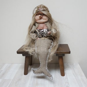 Könnte beinhalten: Eine handgefertigte Puppe mit langen blonden Haaren, die ein Meerjungfrauenkostüm aus Sackleinen mit einem silbernen Paillettenoberteil trägt und auf einem Holzhocker sitzt.