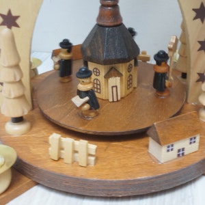 May include: A wooden Christmas pyramid with a church, figures, and trees. The pyramid has four candle holders with white candles.