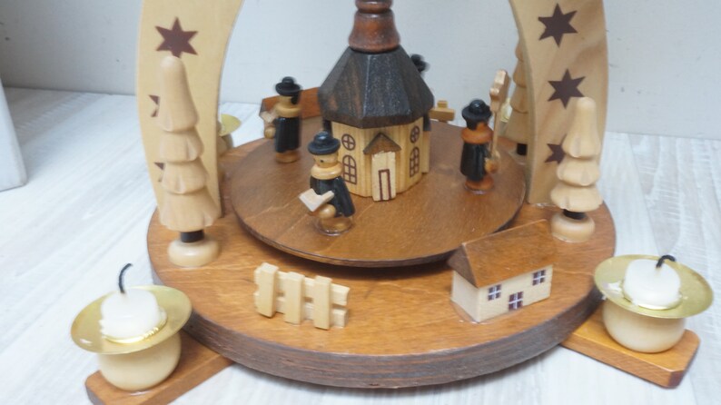 May include: A wooden Christmas pyramid with a church, figures, and trees. The pyramid has four candle holders with white candles.