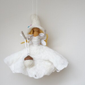 May include: A white felt fairy doll with blonde hair, wearing a white dress and a white hat with blue and pink accents. The fairy is holding a small brown acorn.