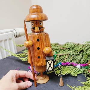 Può includere: Statuina in legno di un uomo con cappello e cappotto, con un bastone e una lanterna. La figura marrone ha dettagli neri e una base rotonda. Accanto c'è un piccolo cono di incenso marrone. Sfondo con vegetazione.