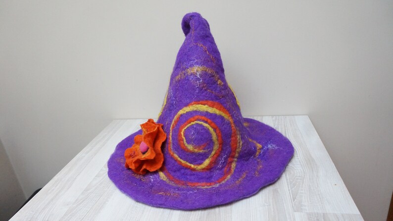 Op de afbeelding: Paarse vilten tovenaarhoed met oranje en gele swirls en een grote oranje vilten bloem.