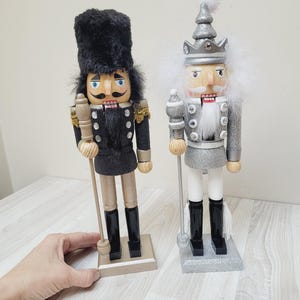 May include: Two wooden nutcracker figurines, one in black and gold with a black fur hat, and the other in silver and white with a silver crown. Both have painted faces and hold a staff.