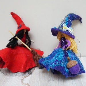 Puede incluir: Dos muñecas de bruja de fieltro, una con un vestido rojo y un sombrero negro, sosteniendo una escoba, y la otra con un vestido azul y un sombrero morado, sosteniendo una pequeña bolsa.