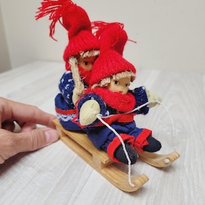 May include: Two small dolls with blonde hair and red knit hats sitting on a wooden sled. The dolls wear blue patterned sweaters, red scarves, and dark pants. A thin white string is attached to the front of the sled, which is made of light-colored wood.