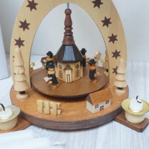 May include: A wooden Christmas pyramid with a church in the center, surrounded by figures and trees. The pyramid has a brown wooden base with three candle holders and a brown wooden arch with stars.