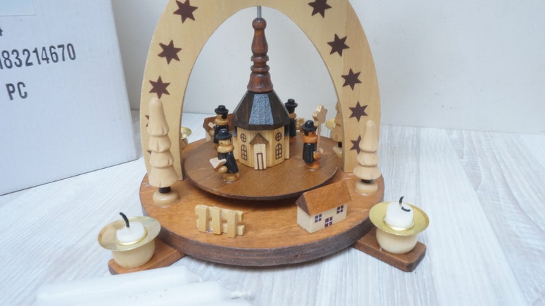 May include: A wooden Christmas pyramid with a church in the center, surrounded by figures and trees. The pyramid has a brown wooden base with three candle holders and a brown wooden arch with stars.