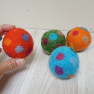 Könnte beinhalten: Vier bunte Filzkugeln mit Punkten. Die Kugeln sind rot, grün, orange und blau. Sie sind aus Wolle und weich und kuschelig.