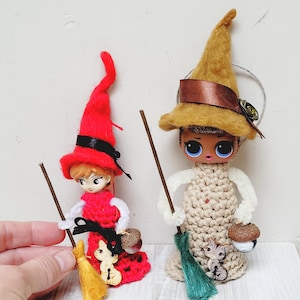 May include: Two handmade witch dolls. One doll wears a red dress and hat with a black ribbon, holding a broom and a small acorn. The other doll has a tan dress and hat with a brown ribbon, holding a broom and an acorn. Both dolls have a cat button.