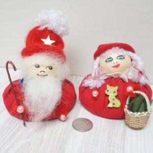 Op de afbeelding: Twee vintage kerstnaaldenkussens, een met een witte baard en een rode muts met een witte ster, de andere met een rode muts en een witte jurk, die een mandje met een groene plant en een gele kat vasthoudt.