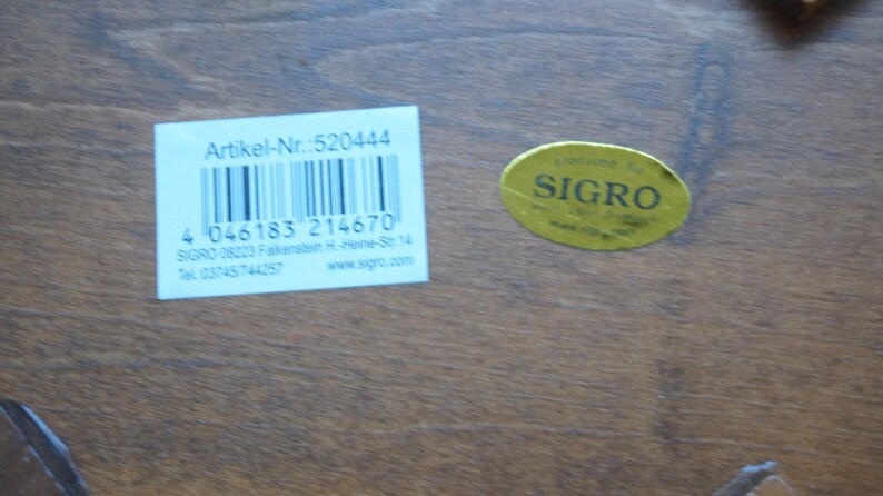 May include: A white label with black text and a barcode. The text reads "Artikel-Nr.:520444", "4 046183 214670", "SIGRO 08223 Falkenstein H-Heine-Str. 14", "Tel: 03745/744257", and "www.sigre.com". A yellow sticker with black text reads "SIGRO".