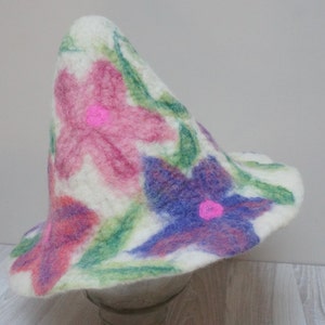 Puede incluir: Un sombrero de fieltro blanco con forma de cono y ala ancha. El sombrero está decorado con flores pintadas a mano en tonos de rosa, morado y verde.