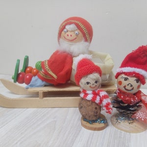May include: Three vintage Christmas decorations, two are red and white and one is a Santa Claus figure on a wooden sleigh. The decorations are made of fabric, wood, and pine cones.