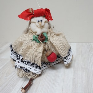 May include: A handmade rag doll with a burlap skirt and a red hat. The doll has a white face with painted features and is holding a red apple.