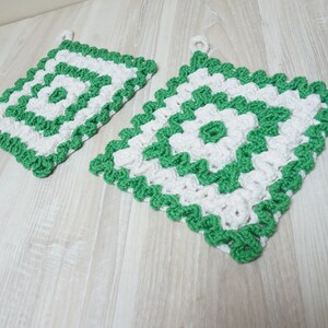 Könnte beinhalten: Zwei gehäkelte Topflappen in einem grün-weißen Muster. Die Topflappen sind quadratisch und haben einen gewellten Rand.
