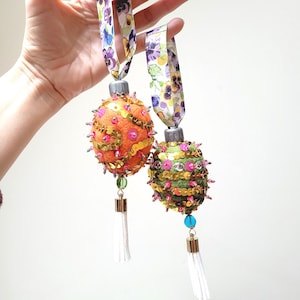 Könnte beinhalten: Zwei handgefertigte Filz-Ornamente mit Pailletten und Perlen. Die Ornamente sind orange und grün mit weißen Quasten.