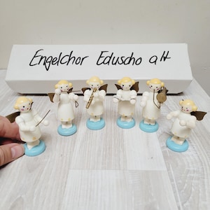 Puede incluir: Un conjunto de seis figuras de ángeles de madera tocando instrumentos musicales. Los ángeles son blancos con cabello amarillo y bases azules y blancas. Los instrumentos incluyen una flauta, una trompeta, un tambor, un violín y una batuta de director. La caja en la que llegaron los ángeles es visible en el fondo con el texto "Engelchor Eduscho a/t".