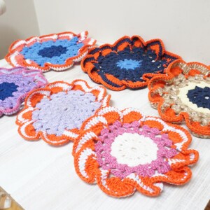Op de afbeelding: Zes gehaakte onderzetters in verschillende kleuren, waaronder oranje, roze, wit, blauw, paars en beige. De onderzetters hebben een gegolfde rand en een centraal ontwerp.
