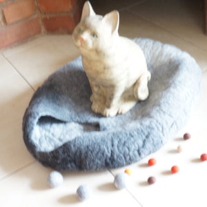 May include: A gray and blue cat bed with a ceramic cat figurine sitting on top. The bed is on a white tiled floor with several small, colorful balls scattered around it.