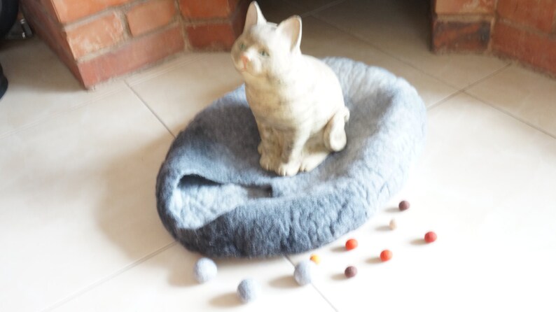 May include: A gray and blue cat bed with a ceramic cat figurine sitting on top. The bed is on a white tiled floor with several small, colorful balls scattered around it.