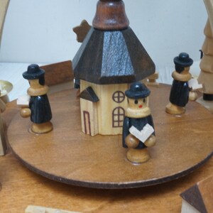 May include: A wooden Christmas pyramid with a brown church in the center, four figures in black coats, and two trees on the sides. The pyramid is decorated with stars.