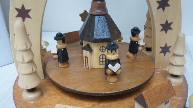 May include: A wooden Christmas pyramid with a brown church in the center, four figures in black coats, and two trees on the sides. The pyramid is decorated with stars.