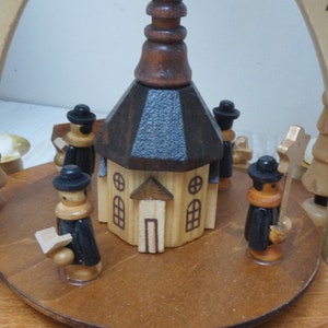 May include: A wooden Christmas pyramid with a church in the center and figures of people around it. The church has a brown roof and a blue roof. The figures are wearing black hats and coats. The pyramid is decorated with stars.